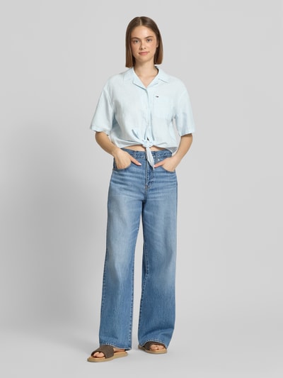 Frauenmodel trägt Regular Fit Leinenbluse aus Leinenmix mit Bindedetail von Tommy Jeans in Hellblau, unifarben, Resortkragen, Logo, kurzarm, Cropped, V-Ausschnitt, Knopfleiste, Ganzkörperansicht von vorne.