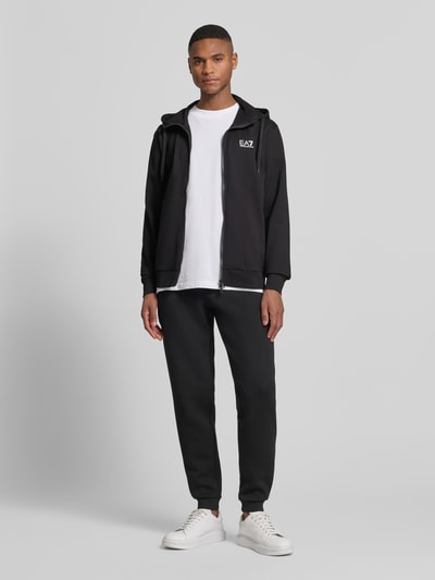 Męski model nosi Slim Fit Sweatjacke mit Kapuze od EA7 Emporio Armani, czarny kolor, mieszanka bawełny, nadruk z logo, wpuszczane kieszenie, elastische wykończenia, tunel w kapturze, stójka, zamek błyskawiczny dwustronny, długość tyłu w rozmiarze M: 69 cm, długość rękawów w rozmiarze M: 69 cm, krój slim fit, widok całej sylwetki z przodu.