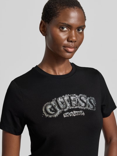 Frauenmodel trägt T-Shirt aus Viskose-Elasthan-Mix mit Logo von Guess, schwarzer Stoff, silbernes Logo, Rundhalsausschnitt, Nahaufnahme Oberkörper.