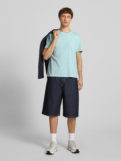 Männermodel trägt T-Shirt aus Baumwoll-Leinen-Mix mit Brusttasche von Superdry in Hellblau, kombiniert mit dunklen Jeansshorts, Ganzkörperansicht von vorne. Regular Fit, Rundhalsausschnitt, Rückenlänge Größe M 66 cm.