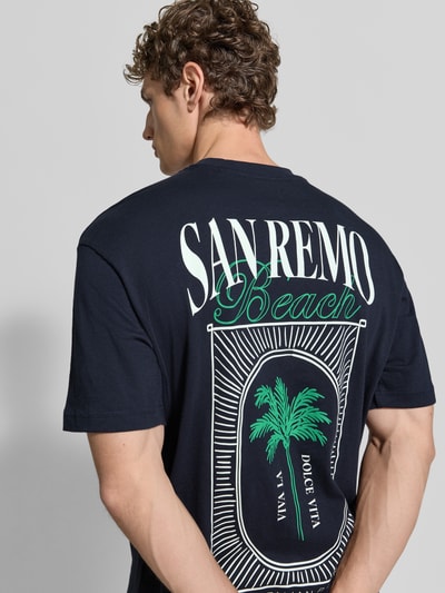 Mężczyzna model nosi T-Shirt aus Baumwolle mit rückseitigem Print od MCNEAL, czarny kolor, krój relaxed fit, nadruk z tyłu, bawełna, okrągły dekolt, długość tyłu w rozmiarze M: 62 cm, całopostaciowy widok z tyłu.