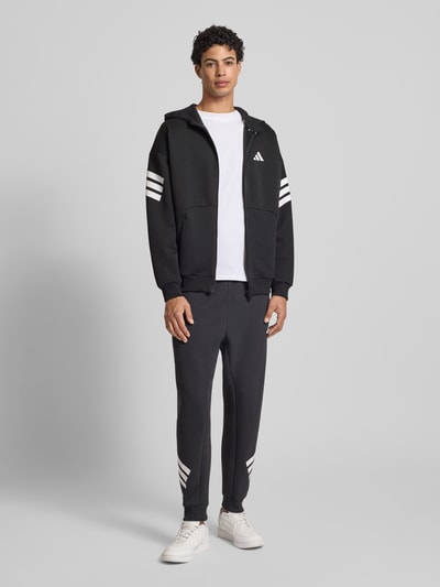 Mannenmodel draagt Regular Fit sweatjack met kangoeroezak van adidas Sportswear, zwarte materiaalmix met katoen, capuchon, ritssluiting, elastische boorden, ruglengte maat M 69 cm, mouwlengte 54 cm, voorkant volledig lichaam.