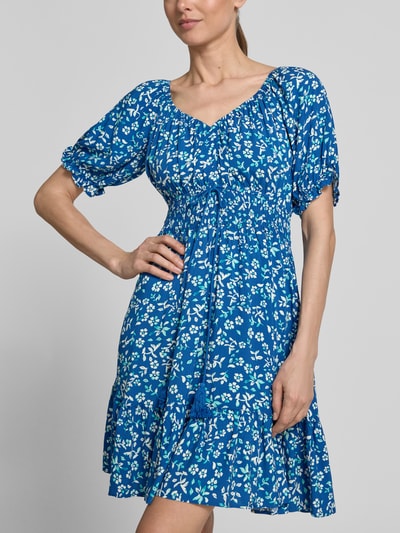 Frauenmodel trägt Minikleid aus Viskose mit Kordelzug und Quasten von Apricot in Blau mit weißem Blumenmuster, Taillierter Schnitt, Rundhalsausschnitt mit Rüschen, Rüschenärmel, Kordelzug, Smok-Details, Rückenlänge bei Größe S 82 cm, Brustweite 34 cm, Ärmellänge 21 cm, Ganzkörperansicht von vorne.