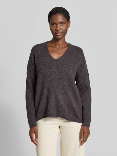 Kobieta nosi sweter z dzianiny z mieszanki wełny z domieszką alpaki, krój regular fit, dekolt w serek, model 'THORA' od MOS MOSH, długość tyłu w rozmiarze S: 82 cm, długość rękawów w rozmiarze S: 46 cm, ciemnoszary kolor, miękka tekstura, widoczne detale splotu, widok całej sylwetki z przodu.