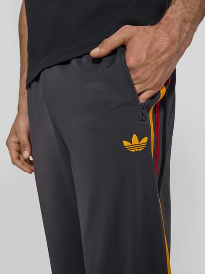 Mężczyzna nosi Relaxed Fit Sweatpants mit Label-Streifen Modell 'FIREBIRD' od adidas Originals, efekt: jednolity kolor, detal z logo, paski z logo, elastyczny ściągacz, tunel, zakończenie nogawek: zamek błyskawiczny, kieszeń zapinana na zamek błyskawiczny, wykończenie materiału: dzianina dresowa, zbliżenie na górną część nogawki z przodu.