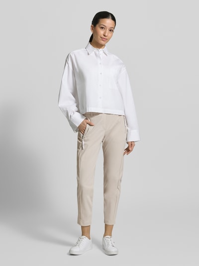 Kobiece modelka nosi Stoffhose in 7/8-Länge mit Stretch-Anteil Modell 'NATINA' od Raffaello Rossi, beżowy kolor, elastyczny materiał, kieszenie na zamek, elastyczny pasek ze szlufkami, krój slim fit, szerokość w rozmiarze 36: 40 cm, długość nogawki od wewnątrz 69 cm, widok całej sylwetki z przodu.
