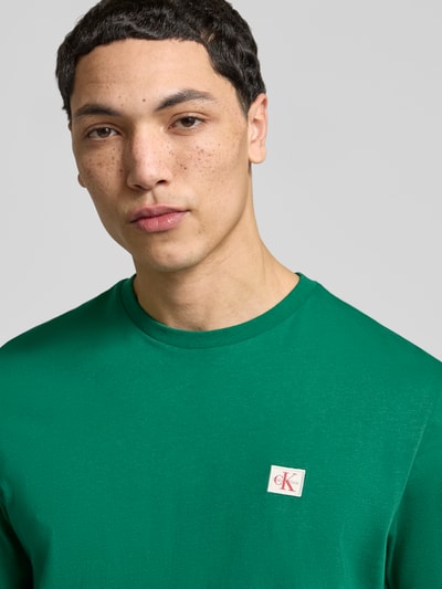 Männermodel trägt T-Shirt mit Label-Badge von Calvin Klein Jeans in Grün, reine Baumwolle, Regular Fit, Nahaufnahme Oberkörper.