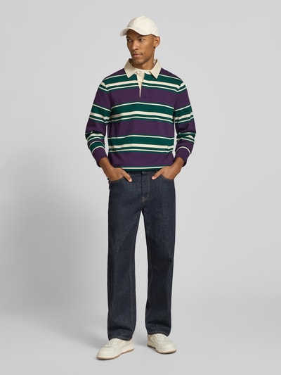 Męski model nosi koszulkę polo Regular Fit Poloshirt aus reiner Baumwolle von GANT w paski zielone, fioletowe i białe, ciemne spodnie jeansowe; widok całej sylwetki z przodu