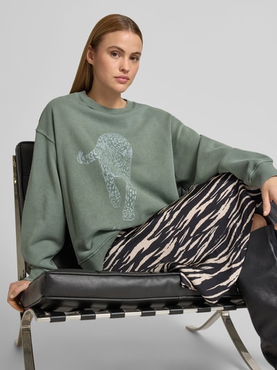 Frauenmodel trägt Oversized Sweatshirt mit Motiv-Print von Lala Berlin in mattem Grün mit Rundhalsausschnitt und elastischen Abschlüssen, reine Baumwolle, weiter Schnitt, Motiv-Print vorn, kombiniert mit gemusterter Hose, Nahaufnahme Oberkörper.