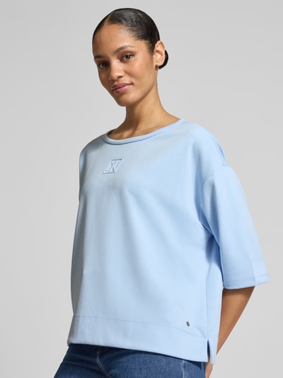 Frauenmodel trägt T-Shirt mit Rundhalsausschnitt und Logo von monari in Hellblau, Relaxed Fit, glatte Oberfläche, Nahaufnahme Oberkörper.