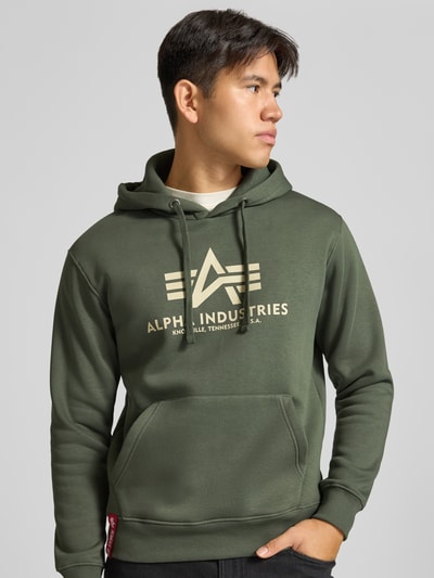 Männermodel trägt Regular Fit Hoodie aus Baumwoll-Mix mit Logo-Print von Alpha Industries in Olivgrün, Logo in Beige, Nahaufnahme Oberkörper. Rückenlänge 70 cm, Ärmellänge 64 cm, Kapuze mit Tunnelzug, elastische Abschlüsse, Label am Saum.
