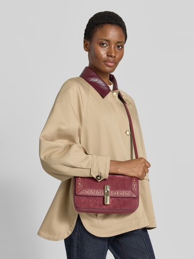 Frauenmodel trägt Crossbody Bag aus echtem Leder Modell 'Iride' von FURLA in Dunkelrot mit Nietenbesatz, glatte Struktur, Nahaufnahme Oberkörper. Hauptfach mit Überschlag, abnehmbarer Schulterriemen, ca. 26x14x6 cm, Riemenlänge 112 cm.
