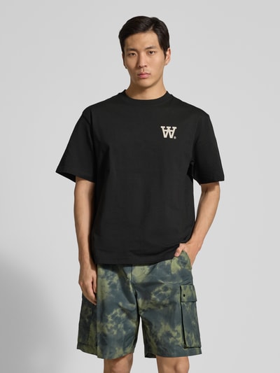 Męski model ma na sobie czarne T-Shirt mit Rundhalsausschnitt Modell 'Liam' od Wood Wood z bawełny, krój regular fit, nadruk z logo, widok całej sylwetki z przodu.