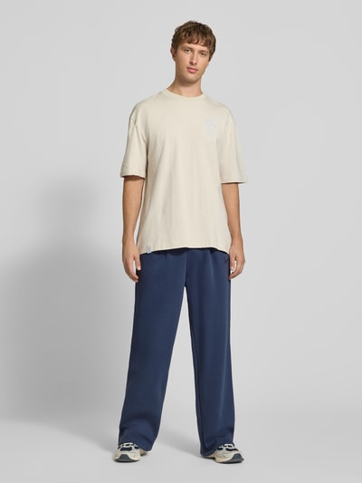 Männermodel trägt marineblaue Sweatpants mit Logo-Stitching von Karl Kani, Wide Fit, Eingrifftaschen, Ganzkörperansicht von vorne.