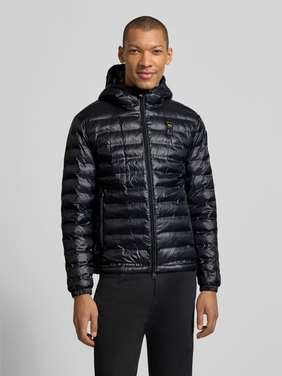 Męski model trägt Kurtka pikowana mit zamek błyskawiczny dwustronny Modell 'Charles' von Blauer Usa w czerni, kaptur, krój regular fit, detal z logo, długość tyłu w rozmiarze M 65 cm, szerokość ramion w rozmiarze M 47 cm, szerokość klatki piersiowej w rozmiarze M 55 cm, długość rękawów w rozmiarze M 65 cm, całość na tle szarego, gładkiego tła. Ganzkörperansicht von vorne.