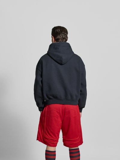 Männermodel trägt Oversized Hoodie aus Baumwolle von RHUDE in Dunkelblau mit Label-Print, Känguru-Tasche, elastische Abschlüsse, Rückenlänge 65 cm, Ganzkörperansicht Rückansicht.