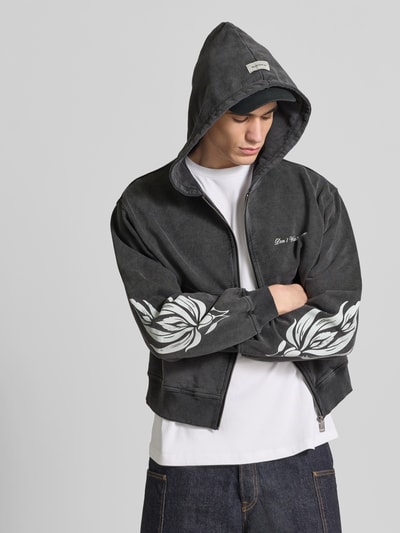 Männermodel trägt Oversized Sweatjacke aus Baumwolle von Don´t Waste Culture in Grau mit floralen Prints und elastischen Abschlüssen, Ganzkörperansicht von vorne. Rückenlänge 63 cm, Schulterbreite 55 cm, Ärmellänge 65 cm, durchgehender Reißverschluss, getrennte Känguru-Tasche.