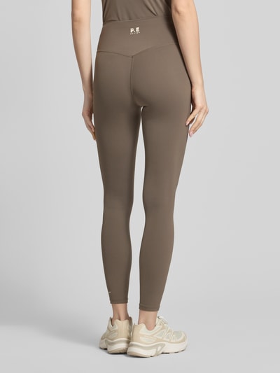 Vrouwenmodel draagt Leggings met elastische tailleband en logo van P.E Nation, bruin, aansluitende high rise pasvorm, stretchmateriaal, logo detail, volledige rugaanzicht.