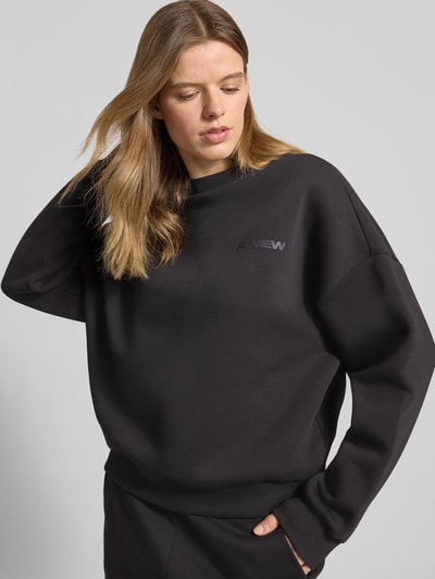 Frauenmodel trägt schwarzen Basic Sweater mit Rundhalsausschnitt und Bündchen am Ärmel von Review, oversized, Logo-Stickerei, Nahaufnahme Oberkörper.