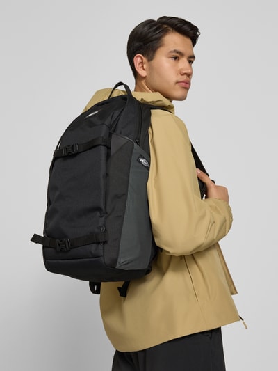 Männermodel trägt schwarzen Rucksack mit Logo-Patch Modell 'ALTACAMA' von Quiksilver, Reißverschlussfächer, Schultergurte, Ganzkörperansicht von vorne.