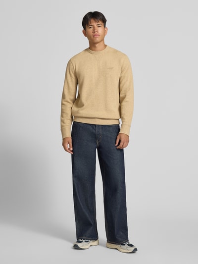 Männermodel trägt Sweatshirt aus Baumwoll-Mix von Superdry in Beige mit Rundhalsausschnitt und elastischen Abschlüssen, lockere Passform, Label-Stitching, kombiniert mit dunkler Jeans und Sneakers, Ganzkörperansicht von vorne.