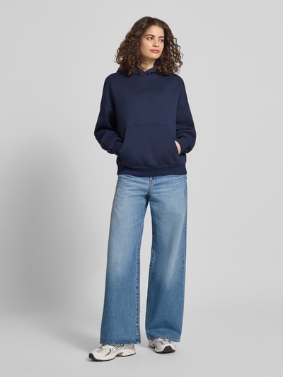 Modelka kobieca ma na sobie Relaxed Fit Sweatshirt mit Logo-Print od Review, bawełniany, granatowy, kieszeń kangurka, krój relaxed fit, widok całej sylwetki z przodu.