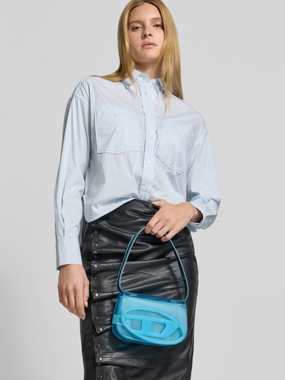 Frauenmodel trägt Shoulder Bag mit Logo-Detail von Diesel in Blau, kombiniert mit hellblauer Bluse und schwarzem Rock, Nahaufnahme Oberkörper.