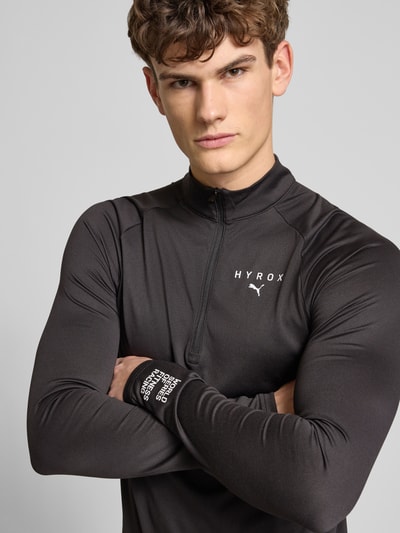 Männermodel trägt PUMA x HYROX - Regular Fit Longsleeve mit Label-Print, Stehkragen, Reißverschluss, Single Jersey, Nahaufnahme Oberkörper.