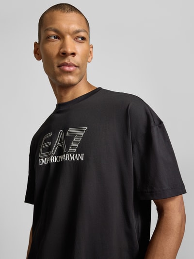 Męski model nosi T-shirt z logo i okrągłym dekoltem od EA7 Emporio Armani z bawełny, krój regular fit, czarny, nadruk logo, długość tyłu 68 cm w rozmiarze M, zbliżenie na górną część ciała.