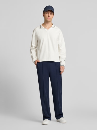 Męski model nosi spodnie dresowe 'Sweatpants mit Längsnähten und Label-Stitching' od CHAMPION z mieszanki bawełny, krój relaxed fit, ciemnoniebieskie, z elastycznym pasem, długość wewnętrzna nogawki w rozmiarze M: 81 cm, całkowity widok z przodu.