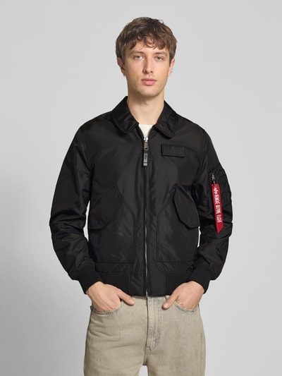 Männermodel trägt Regular Fit Bomberjacke mit Logo-Patch von Alpha Industries, schwarz, Reißverschluss, Umlegekragen, Ganzkörperansicht von vorne.