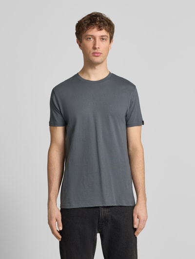 Męski model nosi T-Shirt aus Baumwolle von Alpha Industries w kolorze szarym, prosty krój, okrągły dekolt, detale z logo, długość tyłu w rozmiarze M: 68 cm, widok całej sylwetki z przodu.