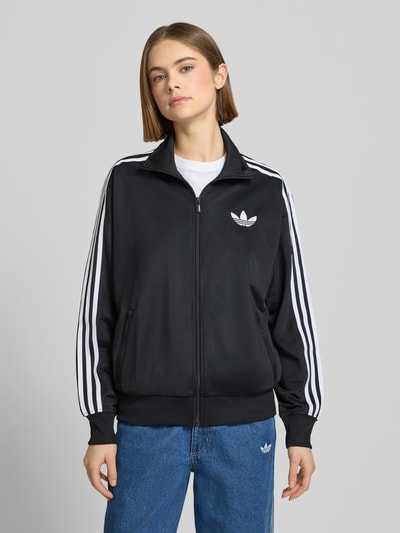 Frauenmodel trägt Sweatjacke mit Stehkragen von adidas Originals in Schwarz mit weißen Streifen, Lockerer-Schnitt, Label-Stitching, Reißverschluss, Reißverschlusstaschen, gerippte Abschlüsse, Ganzkörperansicht von vorne.