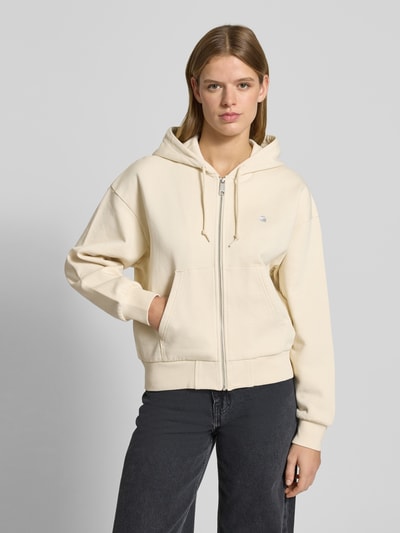 Kobiece model nosi bluzę rozpinaną Sweatjacke mit Känguru-Tasche od Carhartt Work In Progress z bawełny, krój relaxed fit, kaptur, wyhaftowane logo, zamek błyskawiczny, kieszeń kangurka, widok całej sylwetki z przodu.