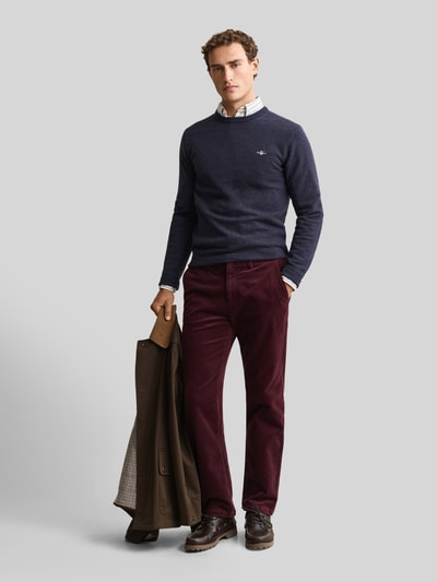 Männermodel trägt Regular Fit Pullover aus reiner Wolle von GANT in Dunkelblau, kombiniert mit bordeauxroser Hose, Ganzkörperansicht von vorne.