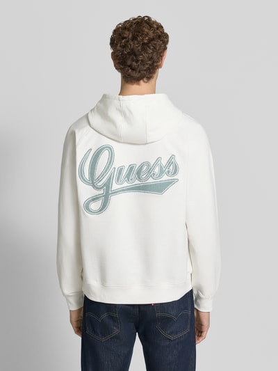 Männermodel trägt Relaxed Fit Hoodie aus Baumwoll-Mix mit Logo-Patch von GUESS, kolor ecru, naszywka z logo, kieszeń kangurka, tunel, krój relaxed fit, długość tyłu w rozmiarze M: 70 cm, długość rękawów w rozmiarze M: 79 cm, mieszanka bawełny, widok całej sylwetki z tyłu.