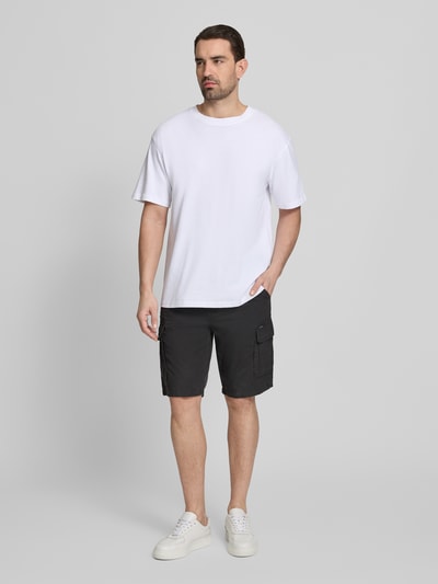 Męski model nosi Regular Fit Bermudas mit Cargo-Taschen von Petrol, czarne bermudy z bawełny, szerokość w rozmiarze M 45 cm, długość wewnętrzna nogawki 27 cm, kieszenie cargo i kieszeń z tyłu, całkowity widok z przodu.