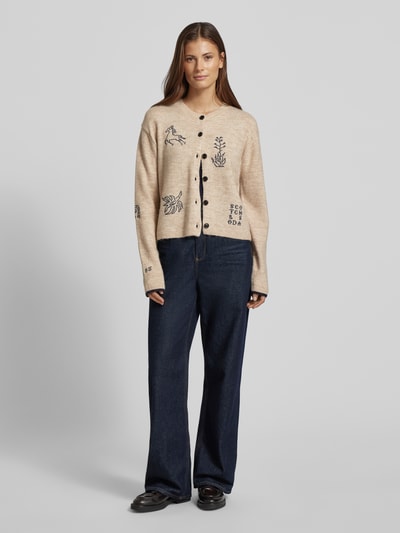 Frauenmodel trägt Strickjacke mit Stitchings und Rundhalsausschnitt von Scotch & Soda, Material-Mix mit Alpaka-Anteil, Regular-Fit, helle Naturfarbe mit schwarzen Stickdetails, Knopfleiste, Rückenlänge 49 cm, Ärmellänge 59 cm, Ganzkörperansicht von vorne.
