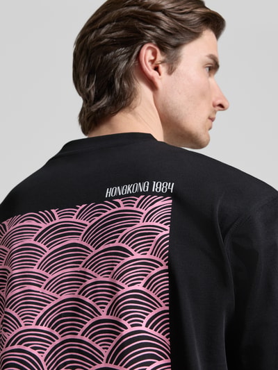 Mężczyznamodel nosi czarny T-shirt z nadrukiem z logo 'T-Shirt mit Label-Print Modell 'Baine'' od Woodbird z bawełny, krój regular fit, okrągły dekolt, długość tyłu w rozmiarze M 67 cm, detal nadruk Hongkong 1984 na plecach, zdjęcie z tyłu.