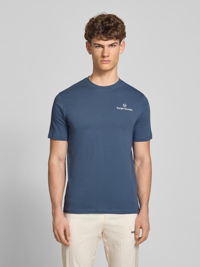Męski model nosi Regular Fit T-Shirt mit Logo-Print von Sergio Tacchini z czystej bawełny, niebieski, fason krój regular fit, widok z przodu, t-shirt z okrągłym dekoltem, długość tyłu w rozmiarze M: 69 cm. Ganzkörperansicht von vorne.