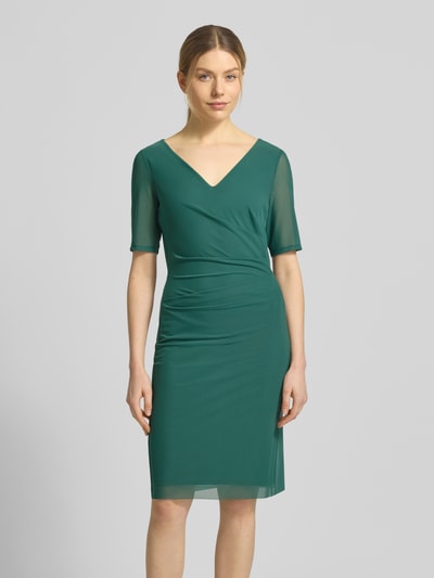 Frauenmodel trägt Regular Fit Midikleid in semitransparentem Design von Vera Mont in Grün, V-Ausschnitt, Kurzarm, Ganzkörperansicht von vorne, elastischer Material-Mix mit blickdichtem Unterrock, Nahtreißverschluss, Rückenlänge 94 cm, Brustweite 37 cm, Ärmellänge 28 cm.