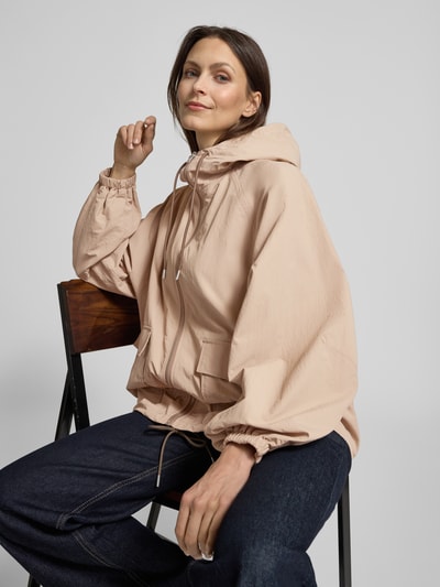 Vrouwenmodel draagt Jacke mit Kapuze und Pattentaschen van Jake*s Casual in beige met capuchon, tunnelkoord, ritssluiting, klepzakken, relaxed fit, ruglengte 66 cm, schouderbreedte 41 cm, borstwijdte 56 cm, mouwlengte 65 cm. Nahaufnahme bovenlichaam.