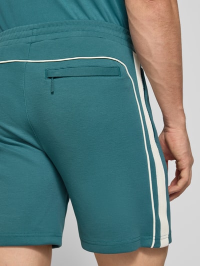 Männermodel trägt Shorts mit Logo-Stitching Modell 'EGEO' von SERGIO TACCHINI in Türkis mit weißen Streifen, Nahaufnahme Rückansicht. Baumwoll-Mix mit Stretch und elastischem Bund, lockerer Schnitt, Detailansicht der seitlichen Eingrifftaschen und Ziertasche.