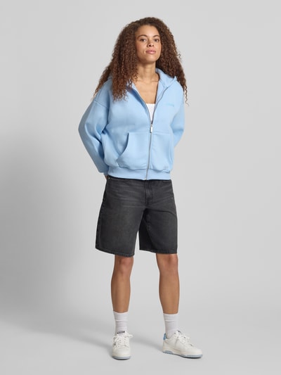 Modelka nosi Relaxed Fit Sweatjacke mit Kapuze od REVIEW, jasnoniebieska bawełna, kieszenie kangurki, zamek, nadruk z logo, widok całej sylwetki z przodu.