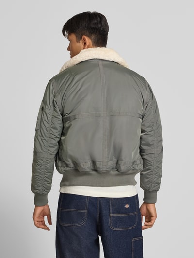 Männermodel trägt Bomberjacke mit Label-Patch und Kentkragen von Alpha Industries in Grau, Teddyfell-Kragen, Ganzkörperansicht Rückansicht. Rückenlänge bei Größe M: 65 cm, Schulterbreite: 45 cm, Brustweite: 56 cm, Ärmellänge: 65 cm, Regular Fit, Reißverschlusstasche am Ärmel, gerippte Abschlüsse.