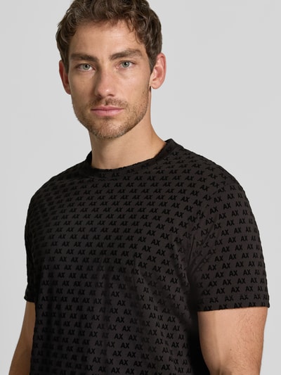 Męski model nosi Regular Fit T-Shirt mit Allover-Print od Armani Exchange, czarny z krótkim rękawem, okrągły dekolt, nadruk na całej powierzchni, Długość tyłu w rozmiarze M: 63 cm, nahaufnahme od przodu.