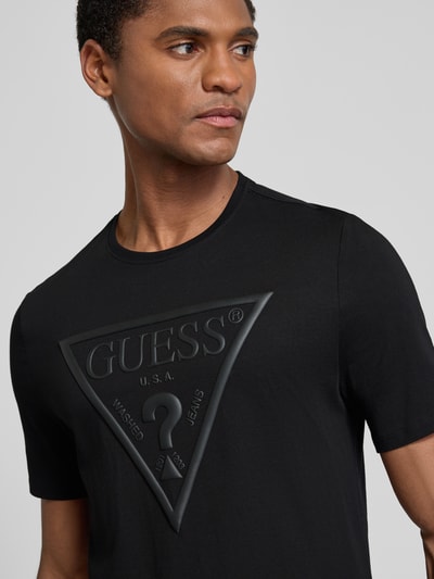 Männermodel trägt T-Shirt mit Logo und Rundhalsausschnitt von GUESS, schwarze Reine Baumwolle, Slim-Fit, Nahaufnahme Oberkörper.