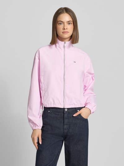 Frauenmodel trägt Relaxed Fit Blouson mit Taillenbändern von Tommy Jeans in Hellrosa, unifarben, Stehkragen, Logo-Stitching, Reißverschluss, lange Ärmel mit elastischen Abschlüssen, seitliche Eingrifftaschen mit Druckknopf, Ganzkörperansicht von vorne.