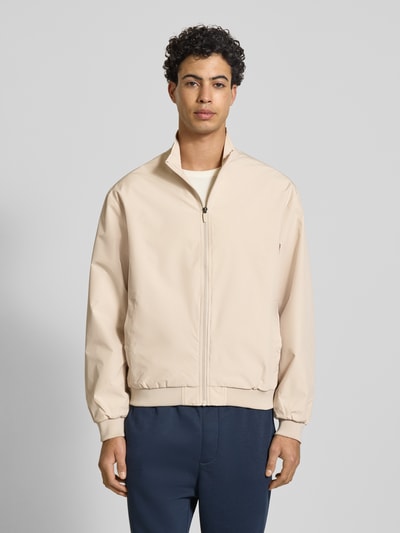 Męski model nosi Regular Fit Kurtka z stójką Model 'Charge' od JACK & JONES, zamek błyskawiczny, wpuszczane kieszenie, elastyczne wykończenia, kolor beżowy; długość tyłu w rozmiarze M: 66 cm, szerokość ramion w rozmiarze M: 49 cm, szerokość klatki piersiowej w rozmiarze M: 50 cm, długość rękawów w rozmiarze M: 64 cm, pełny widok od przodu.