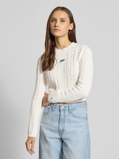 Frauenmodel trägt Slim Fit Strickpullover aus reiner Baumwolle von Tommy Jeans in Optik: Unifarben, Strickoptik, Logo-Detail, Rundhals, Langarm, Materialverarbeitung: Strick, in heller Farbe und gerippte Abschlüsse, Nahaufnahme Oberkörper.